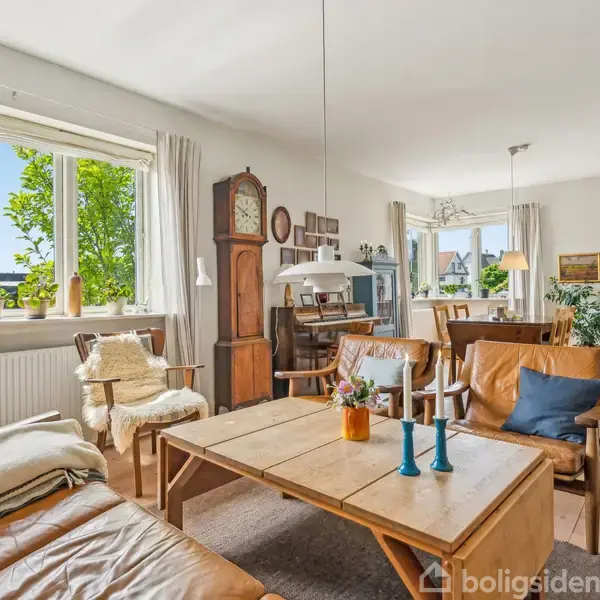 Stue med lædersofa og træbord, dekoreret med blå lysestager og blomster. Rummet er lyst med store vinduer, planter og træmøbler. Vægur og malerier skaber en hyggelig atmosfære.