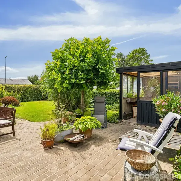 En terrasse med stole og planter er scene for afslapning. En lysthus af glas står ved siden af, omgivet af en velplejet have med grøn græsplæne og buske.