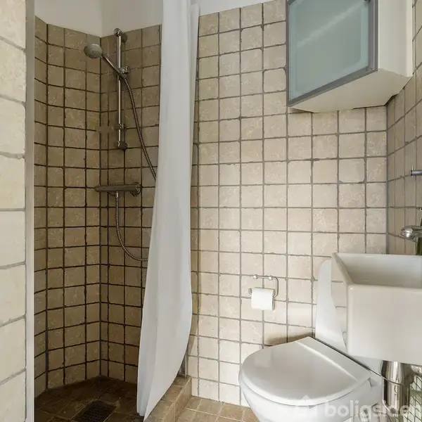 Brusekabine med beige fliser, brusehoved og gardin, placeret i et lille badeværelse med en hvid håndvask, spejlskab og toilet. En sæbedispenser står ved siden af håndvasken.