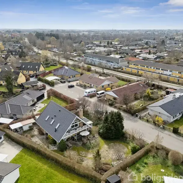 Huse med skrå tage står stille i et boligområde omgivet af grønne haver og veje, med en skolebygning i baggrunden og en parkeret varevogn på en nedre vej.