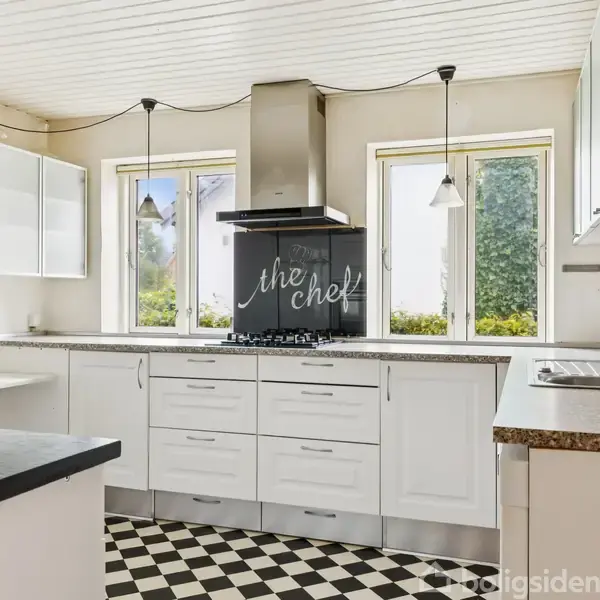 Et moderne køkken med hvide skabe og skakternet gulv. En ovn står midt i rummet under en emhætte. En tavle bag ovnen har teksten “the chef”. Vinduer tillader naturligt lys.