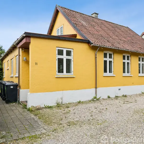 En gulmalet hus med brunrødt tegltag står på en grusbelagt grund, flankeret af affaldsbeholdere og grønne buske. Nummer 11 er synligt på murstensvæggen.