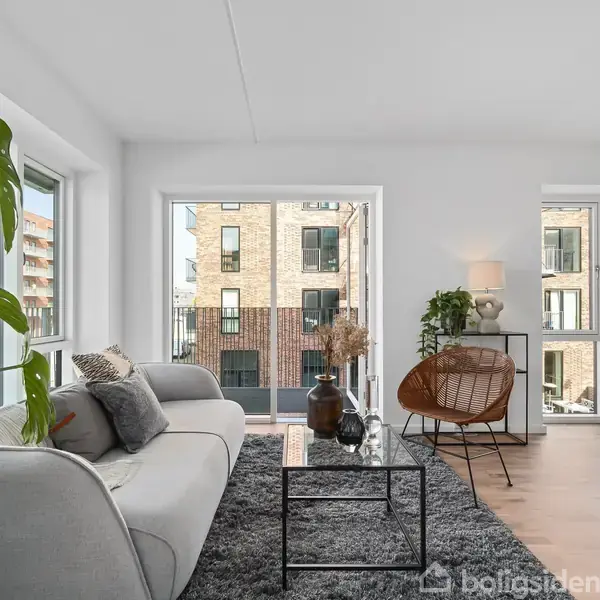 Stue med grå sofa, sort sofabord og grøn plante på tæppe. Store vinduer giver udsigt til nabobygninger. En stol og lampe står ved siden af. Lys og stilfuld indretning.