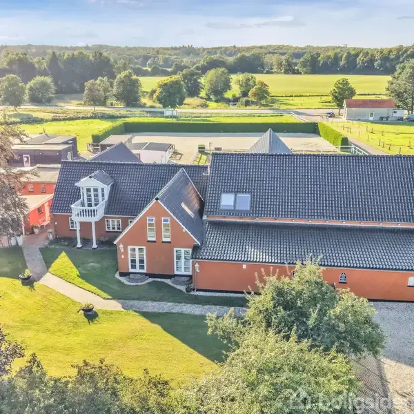 Et rødt hus med sort tag står stille i en grøn have. I baggrunden ses åbne marker og træer under en klar himmel.