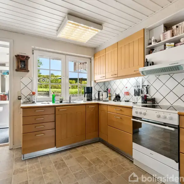 Brunt køkken med hvidevarer mod en væg med fliser; knive hænger på væggen. Til højre ses en døråbning til en stue. Et ur hænger på væggen.