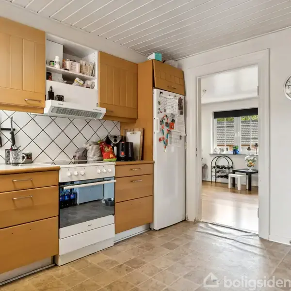 Et køkken med træskabe og fliser. En komfur med hvidevarer og redskaber ovenpå. I baggrunden ses en åben dør til en stue. En vægur viser klokken 10:10.