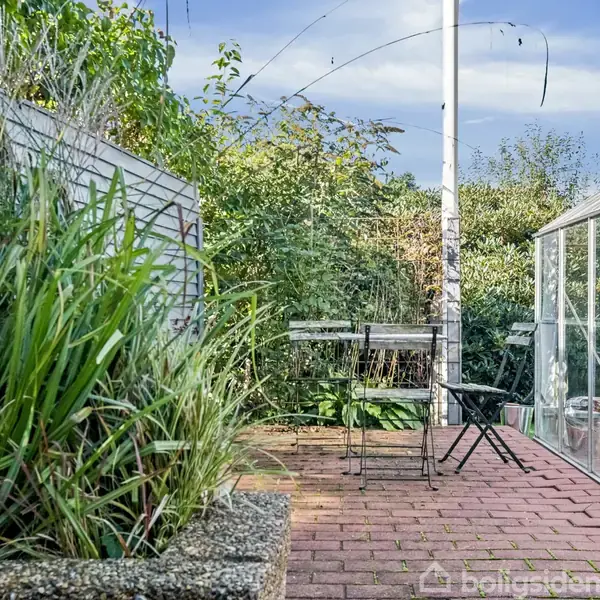 En terrasse med havemøbler, herunder en stol og et bord, står på belægningen ved siden af et drivhus. Omgivet af grøn beplantning og en træhegn i baggrunden.