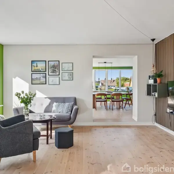 Et spisebord med seks stole står i et lyst rum med grønne vægge. Store vinduer giver udsigt til en frodig have. Blomster og planter pynter rummet.