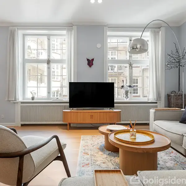 Stue med sofa, lænestol og bord; fjernsyn på skænk. Store vinduer lader lys strømme ind. En lampe med buet stander står ved siden af sofaen. Tresidet bord med dekoration.