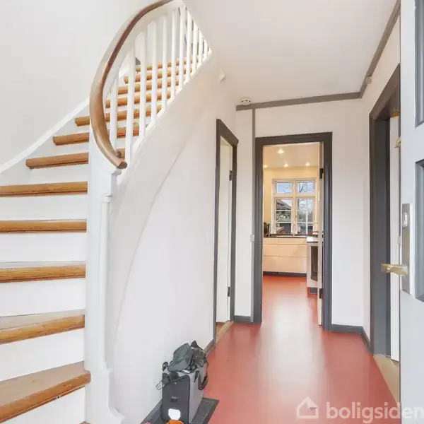 En hvidmalet trappe med trætrin fører opad. Til venstre ligger den og passerer en entre. I baggrunden ses indgangen til et lyst køkkenområde.