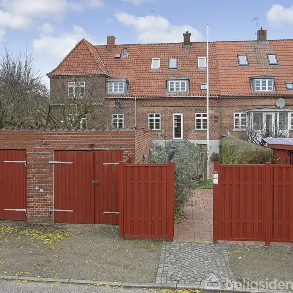 Røde trælåger og en murstensvæg indrammer en havegang foran en række rødmurede rækkehuse med skråtstående tegltag. En flisebelagt sti fører op til bygningerne.