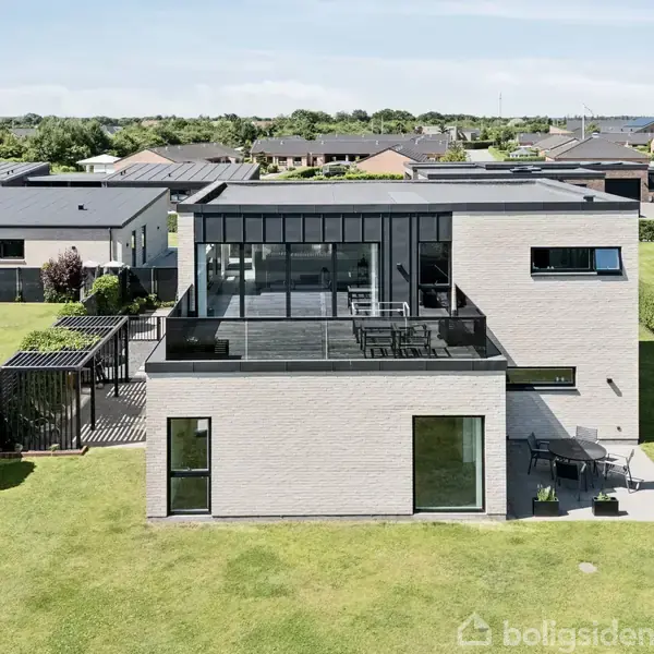 Moderne hus med store vinduer og fladt tag står i en velplejet have. Huset er omgivet af en græsklædt plæne og andre lignende boliger i et forstadskvarter.