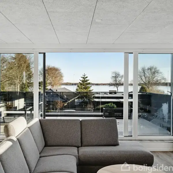 Sofaen står midt i stuen med udsigt til store vinduer, der viser en terrasse og en sø i baggrunden. Væggene er hvide, og en vase står i hjørnet.