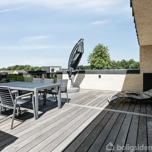 Terrasse med spisebord og stole i moderne design. En stor parasol står ved siden af, og en liggestol er placeret mod væggen. Trægulvet omkranser området, omgivet af grønne træer.