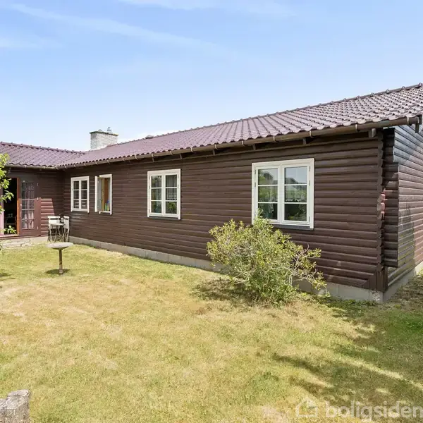 Træhus med mørkebrune bjælker står på en velplejet græsplæne. Omkring huset er der buske og små træer. Himlen er let overskyet, og huset har et rødt tegltag.
