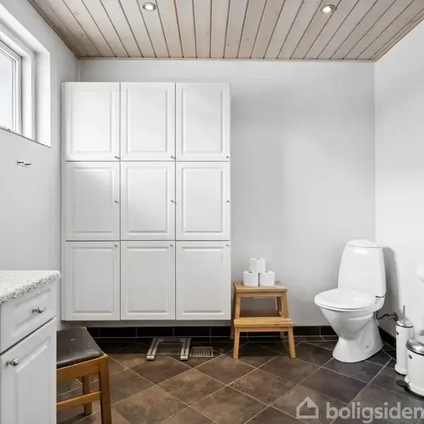 Toilet står i et lyst badeværelse med brunlige fliser. Ved siden af er der et hvidt skab. Ovenpå en skammel er to ruller toiletpapir. Vægbillede: "Reinheimer's Øl".