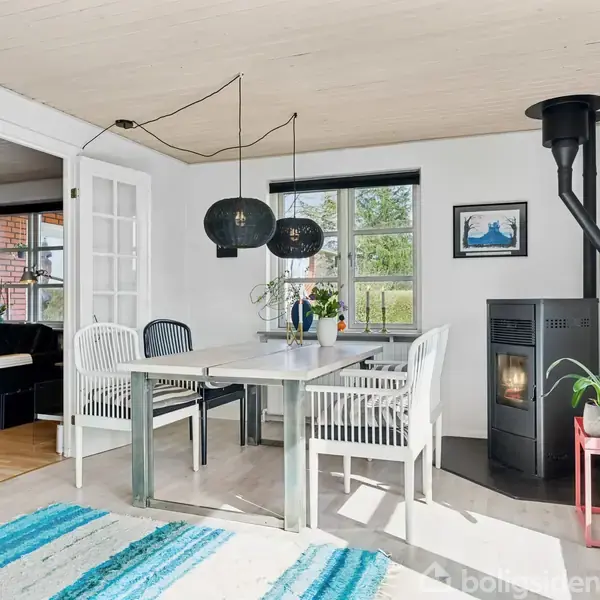 Spisebord med fire stole står i et lyst rum med brændeovn og grønne planter. Store vinduer slipper lys ind, og der hænger sorte lamper over bordet.