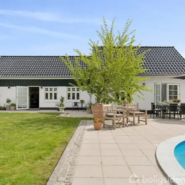 Hus med sort tag omgivet af grøn græsplæne og flisebelagt terrasse. Et træ står ved siden af udendørs spiseborde. Rund swimmingpool ligger til højre under en blå himmel.
