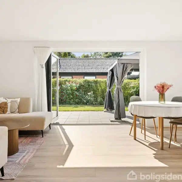 Lys stue med åben glasdør til terrasse. En lys beige sofa står til venstre, og rundt spisebord med blomsterdekoration til højre. Udsigt til grøn have og buske.