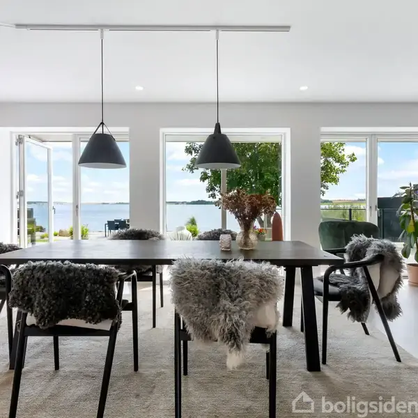 Et spisebord med stole dækket af pels er centralt placeret i en moderne stue. Store vinduer og døre fører til en terrasse med udsigt over vandet udenfor.