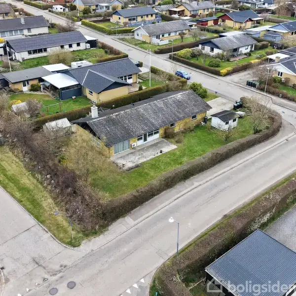 En bolig med en have omgivet af hække ligger i en villavej i et boligkvarter med flere huse, stier og græsarealer omkring.