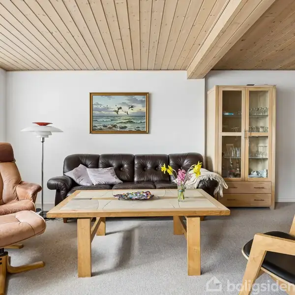 Stue med en sort lædersofa, står foran et stort sofabord med blomster. Til venstre en brun lænestol, til højre en vitrine. Maleri på væggen over sofaen.
