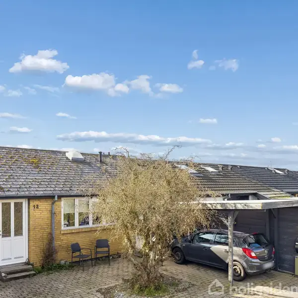 Et enfamiliehus med gule mursten og gråt tegltag står midt i billedet. En grå bil holder parkeret i en carport til højre. Blå himmel med få skyer.
