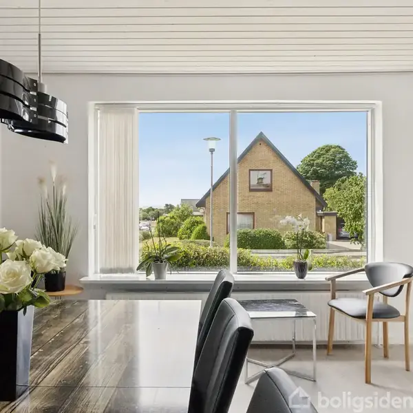 Spisebord med sorte stole står i et lyst rum. Udenfor ses et hus gennem det store vindue. Bordet har en vase med hvide blomster.