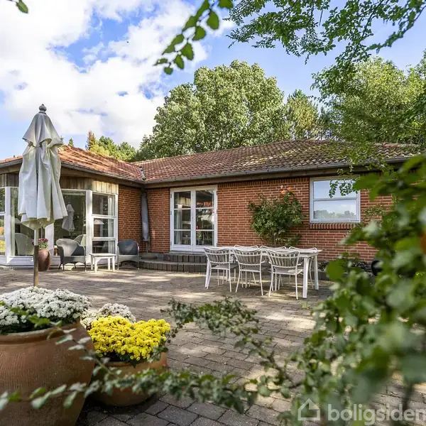 Et rødt murstenshus med en stor terrasse, hvor parasoller og havemøbler står opstillet. Omgivet af masser af grønne træer og buske under en delvist skyet blå himmel.