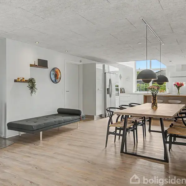 Moderne stue med brændeovn og sort sofa. Spisebord med sorte stole og hængende lamper. Baggrund med åbent køkken, store vinduer, og planteudsmykning giver lys og rummelighed.