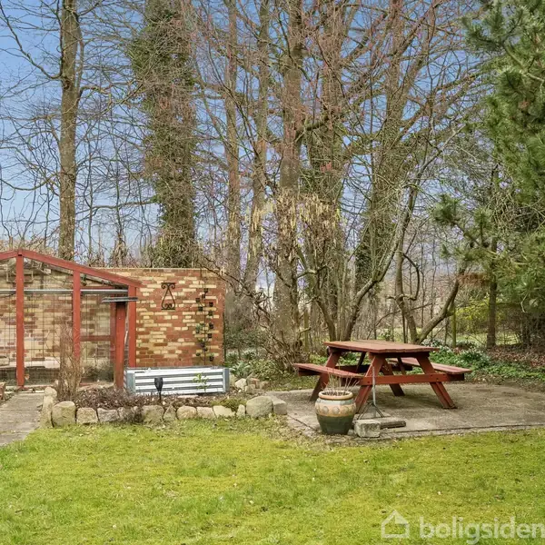 Et havebord står på en terrasse ved siden af et drivhus bygget af mursten og glas. Omgivelserne består af græsplæne, træer og buskads. Text: "boligsiden".