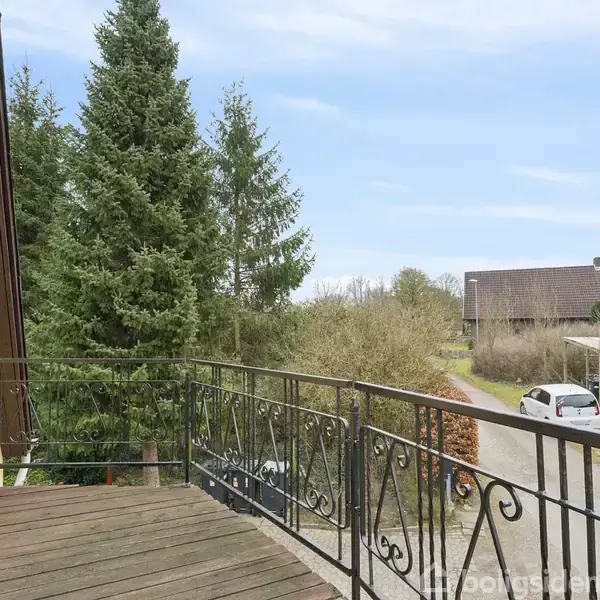 Balkon med trægulv og smedejernsrækværk vender mod en stille vej. En bil er parkeret tæt på, omgivet af store grønne træer og landlige huse i baggrunden.