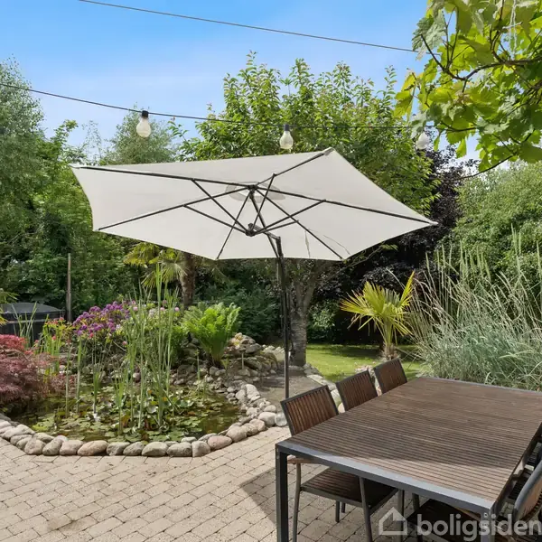 Terrasseområde med havebord og parasol, omgivet af frodig vegetation. En stenbelagt sti fører til en lille dam med blomster og græsser. Lyse lyskæder hænger ovenfor.