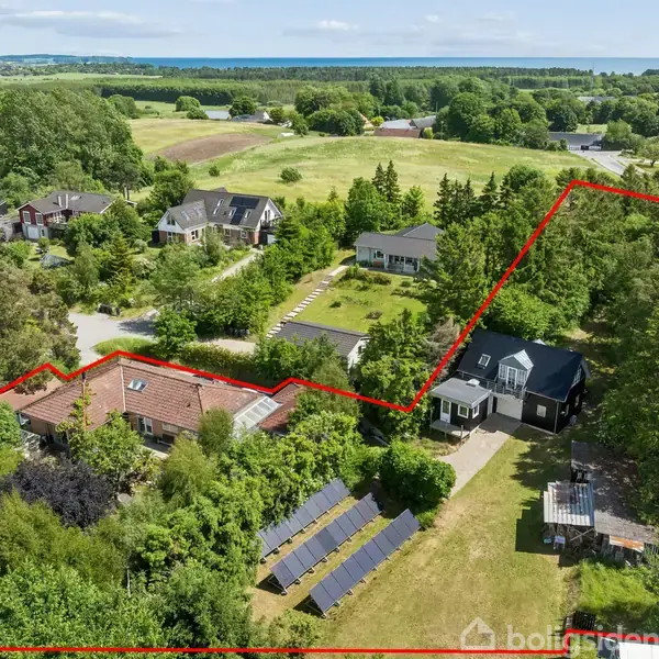 To huse omgivet af frodig vegetation og marker, omkranset af en rød linje. Solpaneler er placeret i haven. En vej løber langs højre side i billedet.