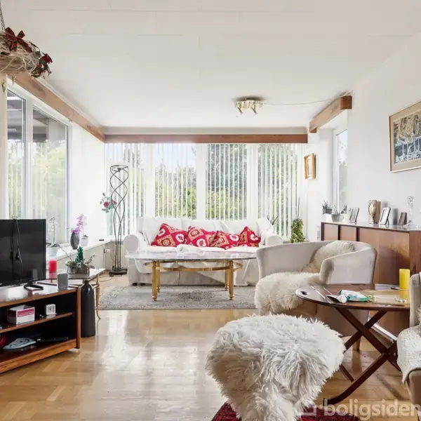 En stue med et fjernsyn på et bord, omgivet af sofaer med fleecetæpper og et sofabord. Store vinduer slipper lys ind, mens en væg har dekorative genstande.