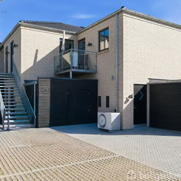 En moderne bygning med lyse mursten, en trappe udenfor og en lille balkon. Bygningen har sorte døre og garageporte i en gårdhave med flisebelagt gulv.