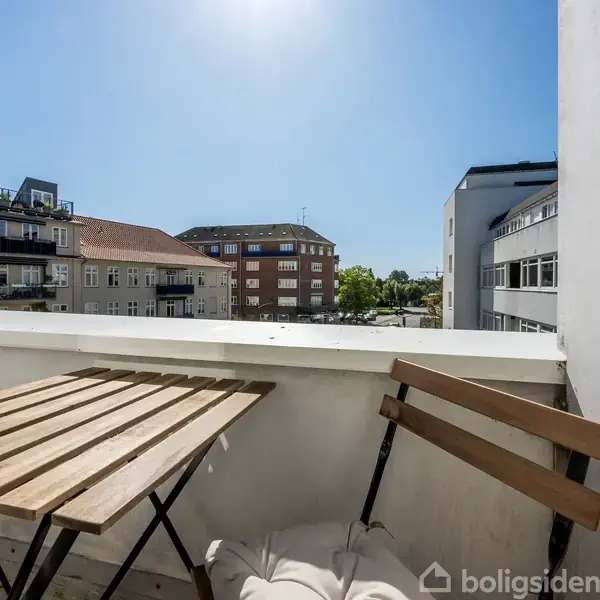 Et træbord med stole står på en altan. Det er solrigt og omgivet af lejlighedsbygninger. Udsigten omfatter en klar blå himmel og grønne træer i det fjerne.