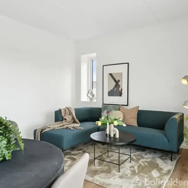 En blå hjørnesofa står i en stue med et tæppe, runde sofaborde og en gulvlampe. Væggene er dekoreret med kunst, mens planter tilføjer grønt til rummet.