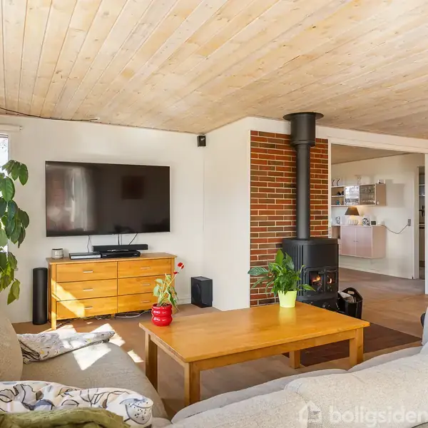 En hyggelig stue med en beige sofa omkring et træbord. En brændeovn står ved en murstensvæg. Et fladskærms-TV hænger på væggen med en skænk nedenunder. Stuen er lys med planter og indgang til køkkenet ses i baggrunden.