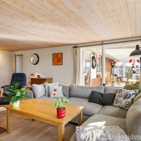 Gråt sofahjørne med puder står i en stue med trægulve og træloft, flankeret af et bord med planter. Stor vinduesparti viser ud til en terrasse. Ingen synlig tekst.