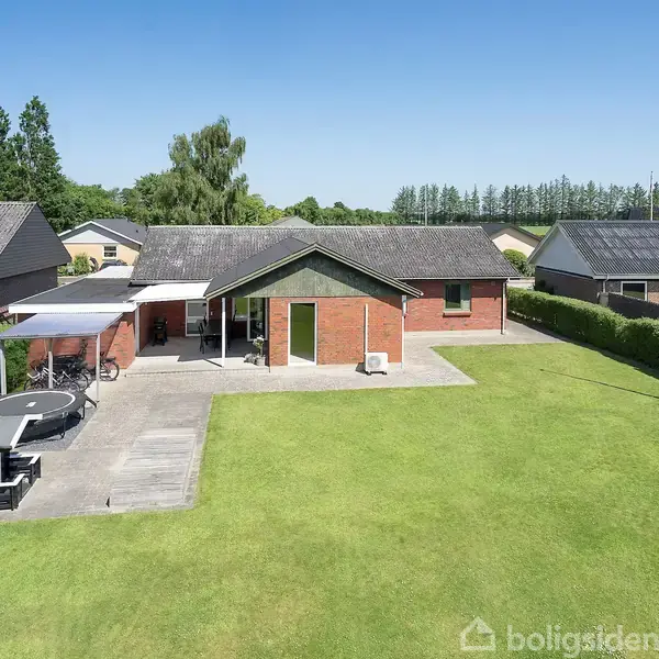 En rød murstensvilla med muret tag. Foran huset er en velplejet græsplæne med en trampolin og en terrasse. Omgivet af hække og andre huse i baggrunden.