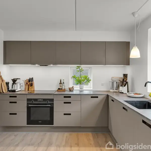 Et moderne køkken med grå skabe og sort bordplade. En ovn er integreret, og et vindue med en plante i potte tilføjer lys. Trægulvet fuldender det stilrene look.