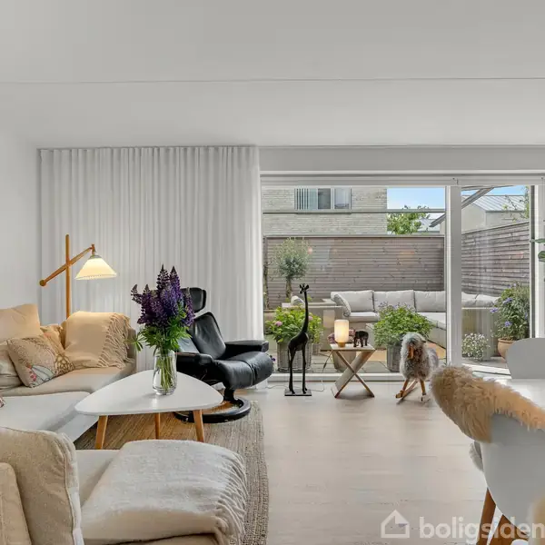 En stue med en beige sofa, der er omkranset af lamper og et maleri på væggen. Rum med store vinduer åbner mod en terrasse med havemøbler og planter.