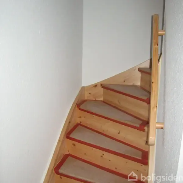 Trætrappe går opad med røde kanter, omgivet af hvide vægge. Trappen drejer i et hjørne, hvilket skaber en cirkulær fornemmelse i det lille rum.