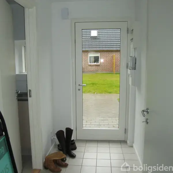 Dør åbner til have, støvler står ved siden af. Udenfor ses en græsplæne og husmur med vindue på en solrig dag.