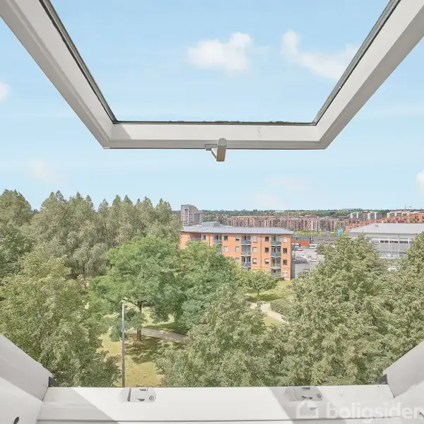 Et åbent vindue giver udsigt over en grøn park med mange træer, mens moderne lejlighedsbygninger kan ses i baggrunden under en klar, blå himmel.