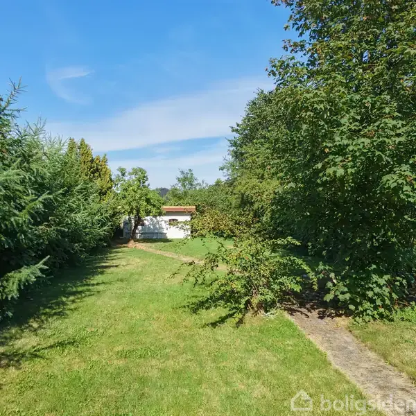 Et hvidt hus står stille for enden af en velholdt, grøn have med en smal sti. Omkring huset er der tætte buske og træer under en klar blå himmel.