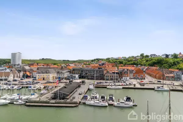 Både ligger til kaj i en havn; bag dem ses en by med røde hustage. Omkringliggende bygninger og en bakke med grøn beplantning skaber en livlig maritim atmosfære.