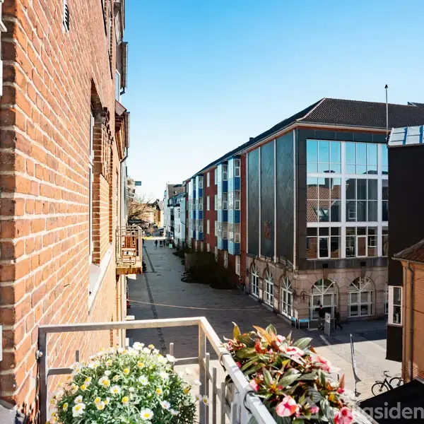 En balkon med blomsterkasser rækker ud fra en murstensbygning. I baggrunden ses en gade flankeret af moderne bygninger med glasfacader under en klar blå himmel.