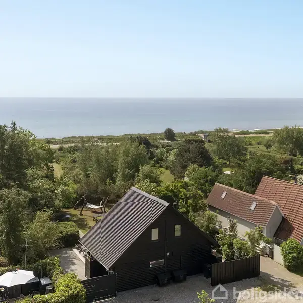 Hus ved vandet omgivet af frodig vegetation. En terrasse med havemøbler er synlig foran huset. Havet og en horisont er i baggrunden, hvilket skaber en fredelig stemning.